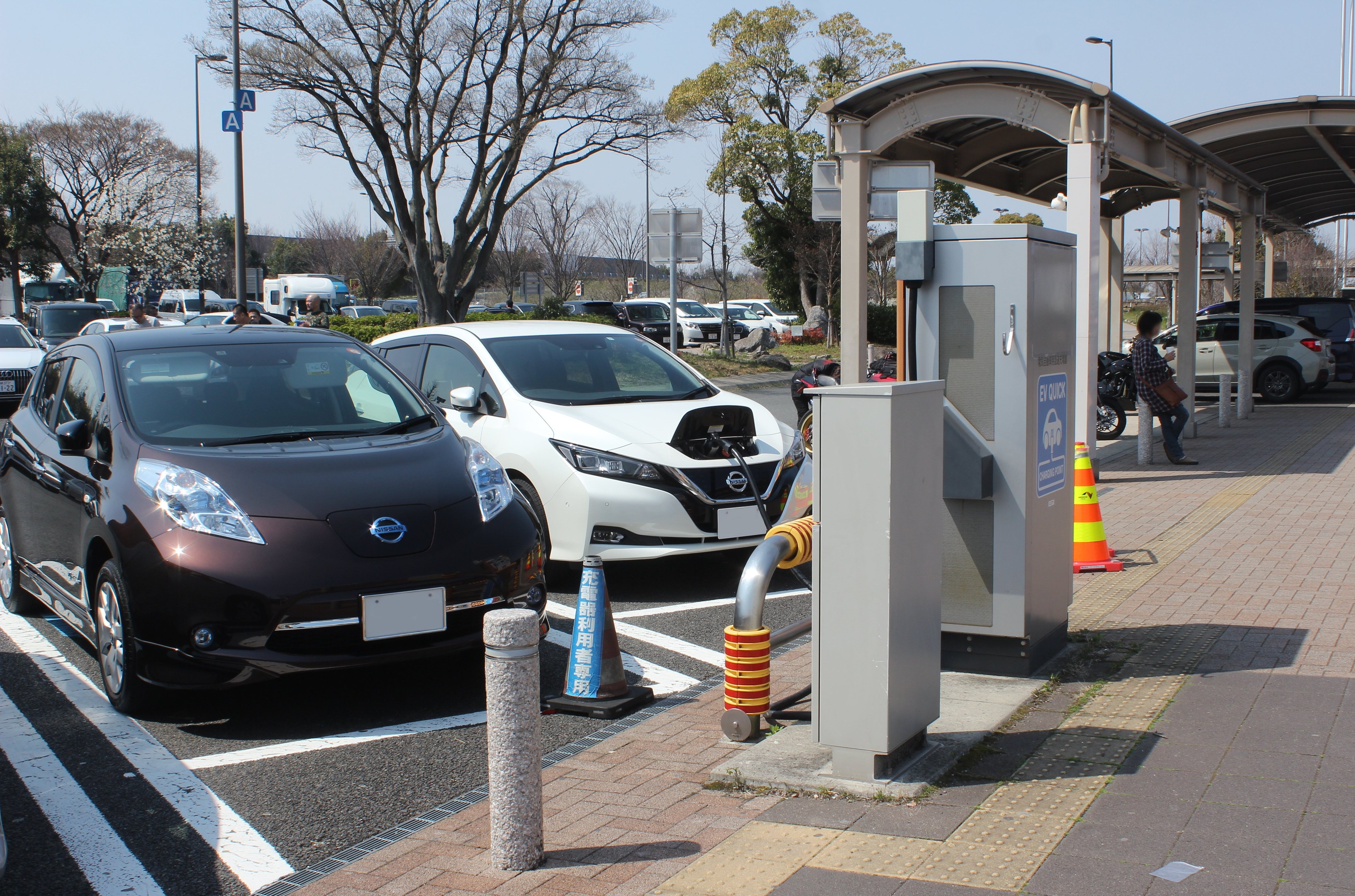 Ladestation für Elektroauto in Japan mit Autos auf der Straße, Verkehrskegel, einer Person auf dem Gehweg, einem Schuppen, Mästen, Lichtern, Schildern, Bäumen, Pflanzen und einem Himmel im Hintergrund.