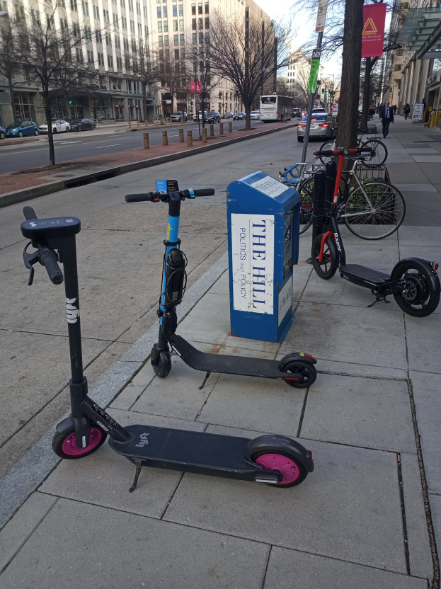 Zwei Elektro-Scooter auf einer Stadtstraße geparkt mit Fahrrädern, einem Müllcontainer und anderen städtischen Elementen im Vordergrund und Fahrzeugen, Bäumen, Gebäuden und Menschen im Hintergrund.