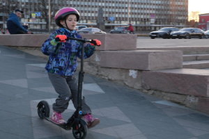 Ein junger Junge mit Helm und Handschuhen fährt auf einem Gehweg mit Treppen, Fahrzeugen, Menschen, Bäumen, Pfosten, Brettern, Gebäuden und einem klaren blauen Himmel im Hintergrund.