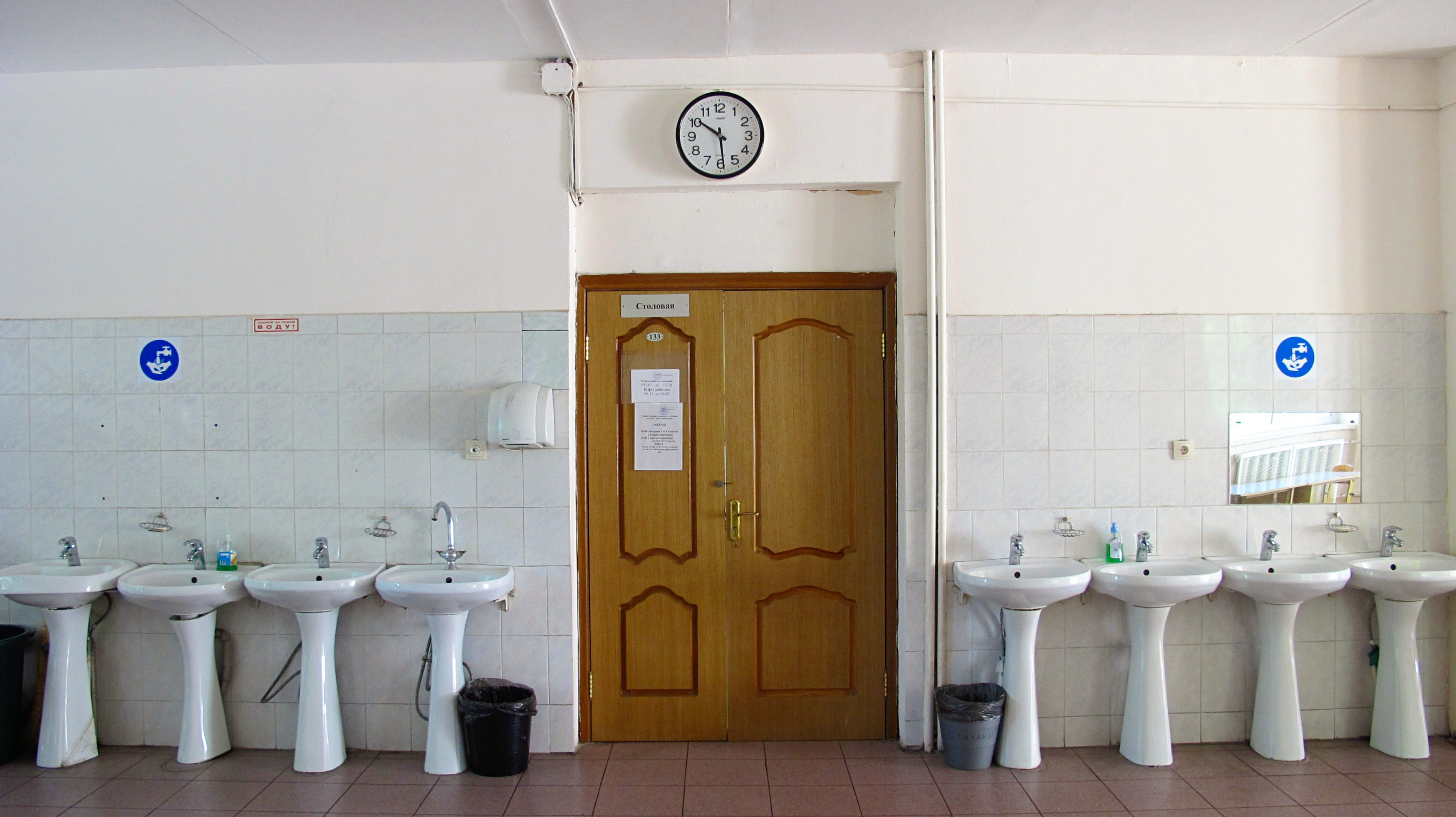 Ein Badezimmer mit mehreren Waschbecken, bodentiefen Müllbehältern, einer Tür im Hintergrund, einer Wanduhr und einem Spiegel auf der rechten Seite.