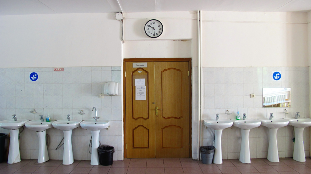 Ein Badezimmer mit mehreren Waschbecken, bodentiefen Müllbehältern, einer Tür im Hintergrund, einer Wanduhr und einem Spiegel auf der rechten Seite.