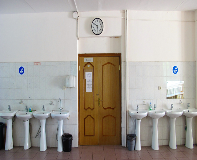 Ein Badezimmer mit mehreren Waschbecken, bodentiefen Müllbehältern, einer Tür im Hintergrund, einer Wanduhr und einem Spiegel auf der rechten Seite.