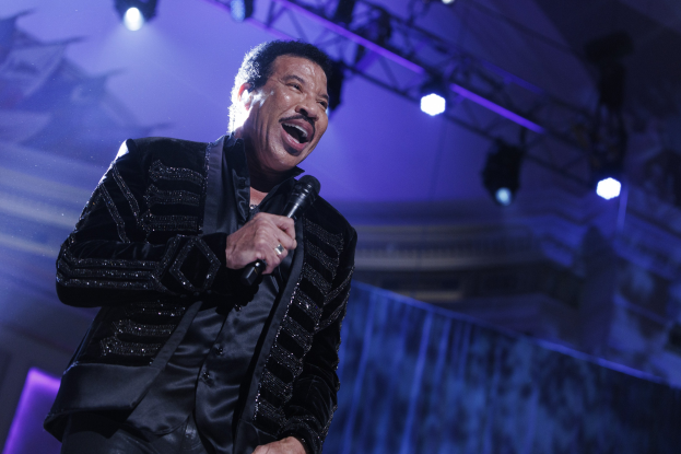 Lionel Richie performs auf den Grammys, trägt ein schwarzes Kleid und hält ein Mikrofon, während helle Bühnenlichter eine dramatische Atmosphäre schaffen.