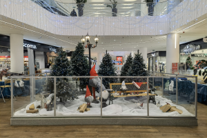 Ein festlich geschmückter Weihnachtsmarkt in einem Einkaufszentrum mit geschmückten Tannenbäumen, Schneeflocken, Sternen und Efeublättern sowie Menschen und Einkaufszentrum-Elementen im Hintergrund.