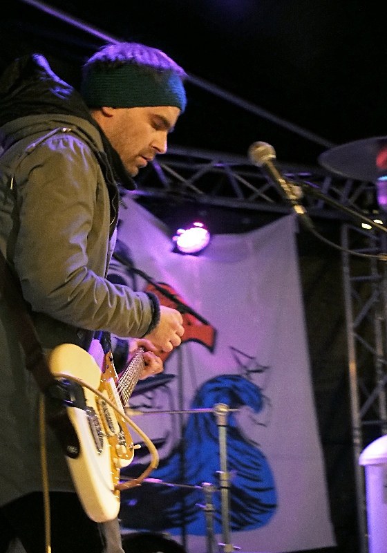 Ein Mann in einer Jacke und einer Mütze spielt eine E-Gitarre auf der Bühne, mit einem Mikrofon vorne, umgeben von Scheinwerfern, einem Banner und einem Schlagzeug zu seiner Rechten.