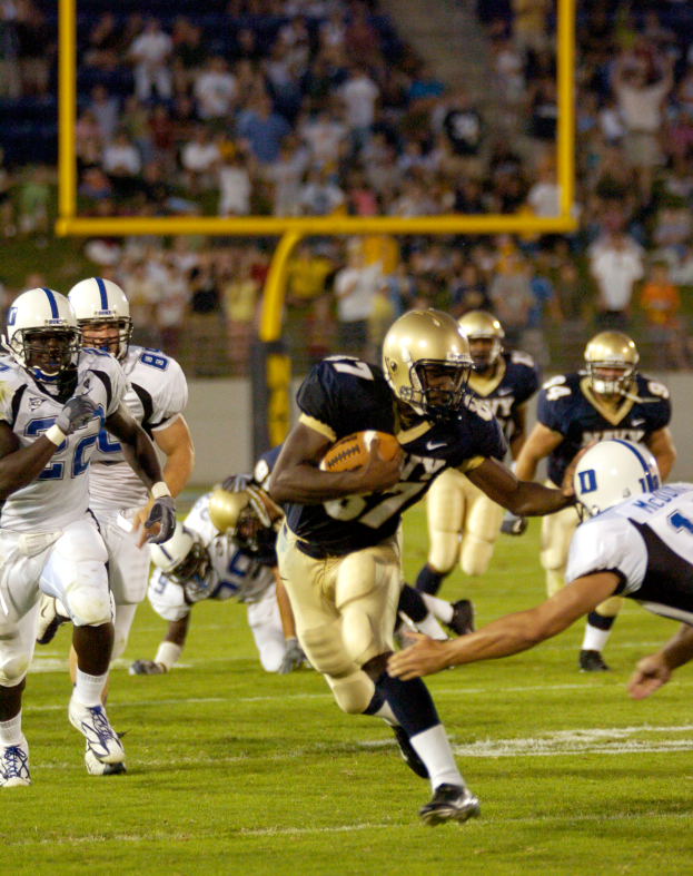 Ein Fussballspieler in einem Sportoutfit und Helm rennt mit dem Ball, ├╝mgeben von anderen Spielern, mit einem unscharfen Hintergrund und Text unten auf dem Bild.