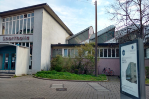 Ein großes Gebäude mit einem Schild davor, umgeben von Bäumen, Pflanzen, Gras, Fahrrädern und einem Pfahl, unter einem bewölkten Himmel, identifiziert als das Sportballe, soll im Jahr 2020 eröffnet werden.