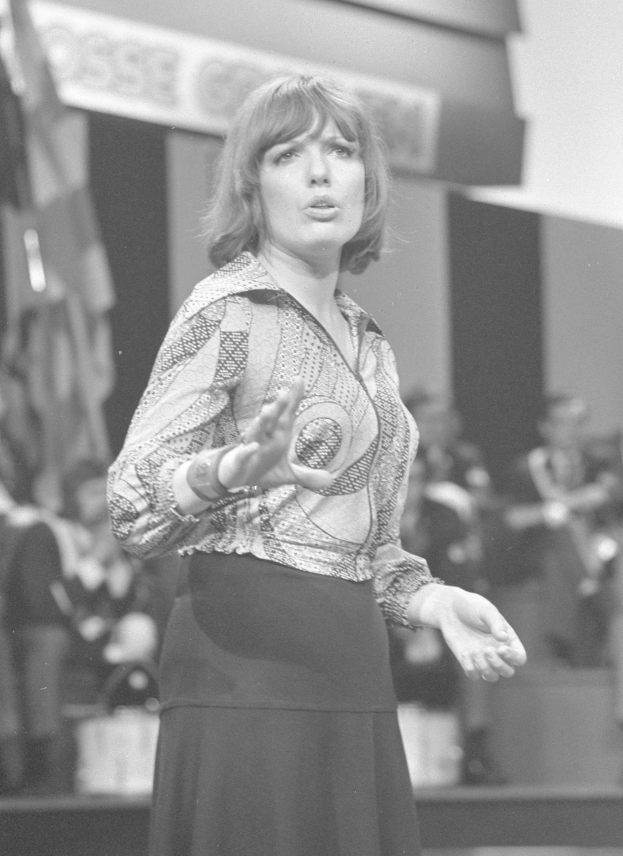 Eine Frau in einem Schwarz-Weiß-Foto steht auf einer Bühne, trägt ein weißes Hemd und einen schwarzen Rock, hat ihr Haar zu einem Dutt gebunden und einen ernsten Gesichtsausdruck, umgeben von einer Gruppe von Menschen und einer Wand im Hintergrund.