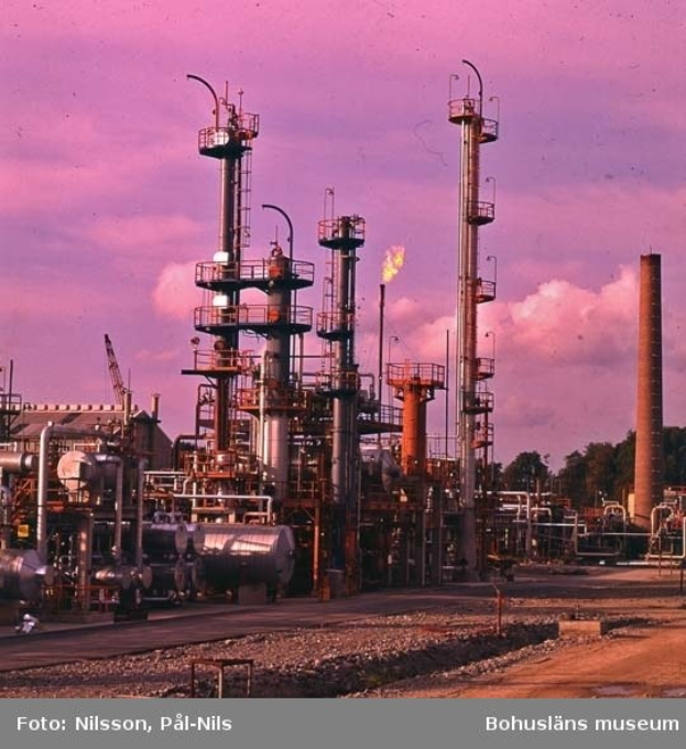 Industrielle Ölfabrik mit ausgedehnten Rohrleitungen, Metallstrukturen, Containern, Bäumen, einem Gebäude, einem Kran und einem bewölkten Himmel, mit Text unten.