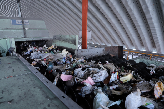 Großer Müllberg auf einem Förderband in einer Recyclinganlage mit einer Maschine im Vordergrund und einem Metalldach darüber.