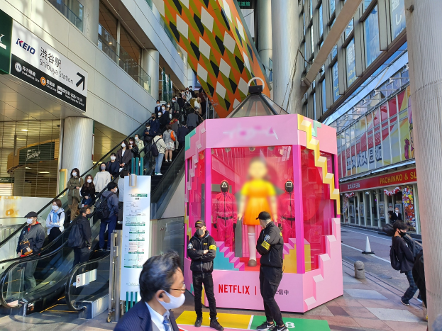 Eine Gruppe von Menschen steht vor einem Gebäude mit Glaswänden, Säulen und Texttafeln, einige tragen Masken, während andere eine Rolltreppe verwenden; ein pinker Kasten in der Mitte zeigt Schaufensterpuppen und trägt die Aufschrift "Netflix's new Netflix Booth in Tokyo."