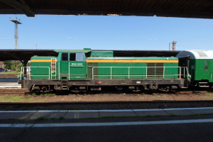 Grüne Diesel-Elektrolokomotive auf Eisenbahnschienen unter einer Brücke, umgeben von Steinen, Gras, einem Weg, Bäumen, Strommasten mit Drähten, einem Turm, Fahrzeugen und einem bewölkten Himmel.