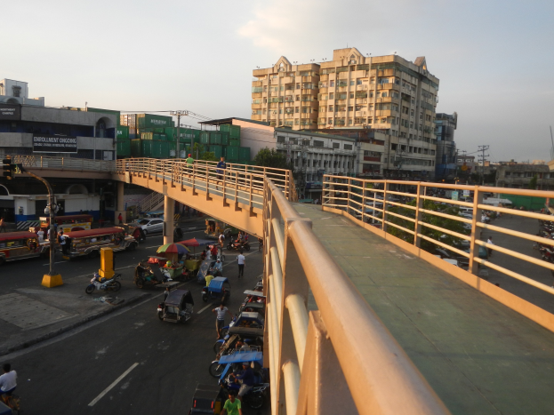 Eine Brücke mit Geländern überspannt eine belebte Straße mit vielen Fahrzeugen, Fußgängern mit Schirmen und anderen Gegenständen, mit Gebäuden, Bäumen, Pfählen und einem klaren blauen Himmel im Hintergrund.