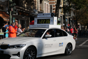 Ein weißer BMW mit einer Uhr auf dem Dach fährt eine Straße entlang, mit einer Person im Auto und einer Gruppe von Menschen am Straßenrand, während im Hintergrund Gebäude, Bäume und Ampeln zu sehen sind.