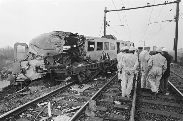 Ein Schwarz-Weiß-Bild einer Zugkollision mit Menschen in Helmen drumherum, Strommasten, Bäume, Gebäude und einen klaren Himmel im Hintergrund.