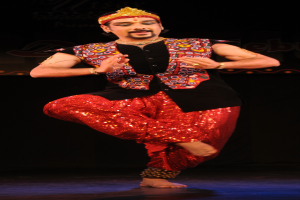 Ein Mann in roter und schwarzer Kleidung tanzt auf einer Bühne mit dunklem Hintergrund, sein Gesicht ist zu sehen, die Augen sind geschlossen und der Mund leicht geöffnet.