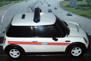 Weißes Polizeiauto mit "Polizei"-Aufschrift gefolgt von mehreren Fahrzeugen auf einer Straße, flankiert von Gras und Bäumen, vor einer Stadt.