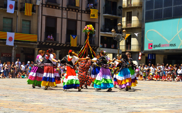 Eine Gruppe von Frauen in bunten Kleidern tanzt auf der Straße, umgeben von einer Menschenmenge, mit Gebäuden, Geländern, Balkonen, Fahnen, einem Laternenmast und einer Tafel mit Schrift im Hintergrund.