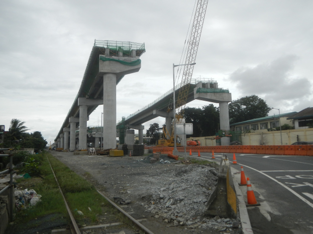 Baustelle mit einer Brücke im Hintergrund, eine Straße mit Absperrkegeln auf der rechten Seite, Steine und Gras auf dem Boden, eine Bahnschiene auf der linken Seite, Bäume und Gebäude auf beiden Seiten der Straße und ein bewölkter Himmel.