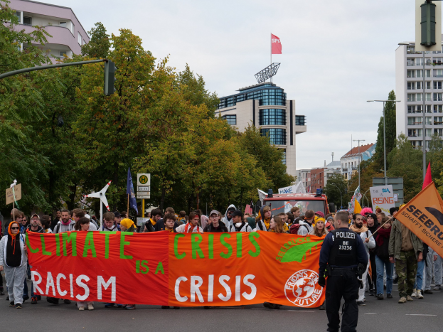 Eine Gruppe von Menschen marschiert mit einem Banner, auf dem "Klima-Krise ist eine Krise" steht, eine Straße entlang mit Bäumen, Laternenmasten und Gebäuden am Straßenrand unter einem klaren blauen Himmel.