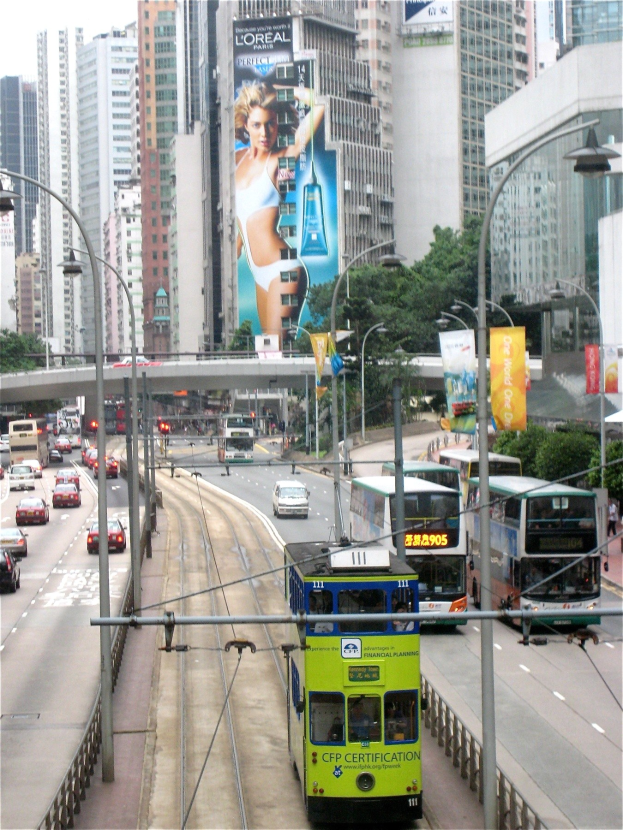 Eine belebte Stra├če mit Doppeldeckerbussen, Autos und anderen Fahrzeugen unter einem klaren blauen Himmel, mit hohen Geb├Ąuden, Laternen, Schildern, Bannern, B├Ąumen und einer Brücke im Hintergrund, mit Text unten.