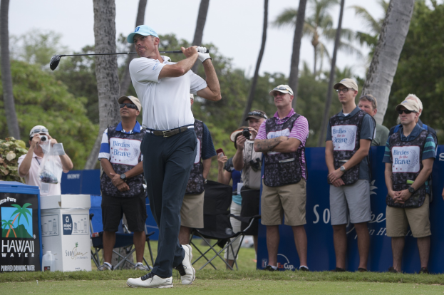 Ein Mann in einem weißen T-Shirt, schwarzen Hosen und einer blauen Mütze schwingt einen Golfschläger auf einem saftig grünen Feld, mit Zuschauern und Kameras dahinter, vor einem Wald und einem klaren blauen Himmel.