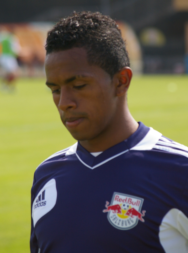 Ein junger Mann in einem blauen T-Shirt steht auf einem Fußballfeld, der als Flügelspieler mit kurzen dunklen Haaren und entschlossener Miene gegen einen unscharfen Hintergrund erscheint.