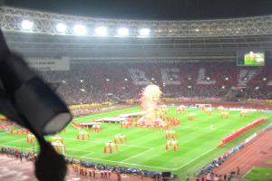 Ein Stadion mit Flutlicht, Eisengittern, Zuschauern, Menschen auf dem Boden, Werbetafeln, Anzeigetafeln, einer Überwachungskamera, Fahnen und Dekorationen.