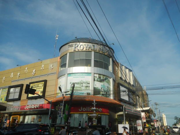 Eine belebte Stadtstraße mit fahrenden Autos, Fußgängern auf dem Gehweg und einem großen Gebäude mit der Aufschrift "Königliches Einkaufszentrum" im Hintergrund, umgeben von parkenden Autos, Bäumen, Strommasten und Drähten unter einem Himmel.