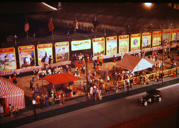 Ein altes Nachtkarnevalsbild mit Menschen, einem Auto, Masten mit Fahnen, einem Zaun, einem Zelt, Brettern mit Bildern und Text sowie einer Straßle.