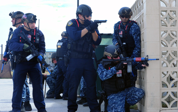 Gruppe von Polizisten in Helmen und Uniformen auf einer Brücke stehend, bewaffnet, mit einer Wand auf der rechten Seite und Gebäuden, Bäumen und Himmel im Hintergrund.