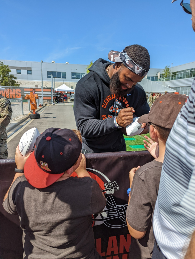 Cleveland Browns Wide Receiver Odell Beckham Jr. signiert Autogramme für Fans auf dem Trainingsgelände, umgeben von Menschen mit Mützen, einem Banner und Gebäuden im Hintergrund bei einem bewölkten Himmel.