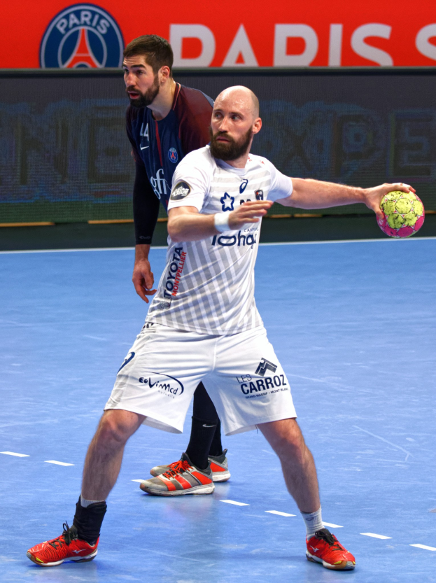 Zwei Männer spielen Handball auf einem Court, einer hält den Ball, mit einem Hintergrundschild, auf dem "Paris Saint-Germain vs Paris Saint-Germain" steht.