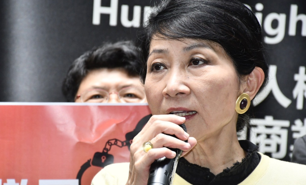 Frau in schwarzem Kleid mit Mikrofon, die vor einer Menge spricht und ein Banner mit der Aufschrift \'Hong Kong Chief Executive Carrie Lam\' hinter sich hält, mit entschlossener Miene und selbstbewusster Haltung.