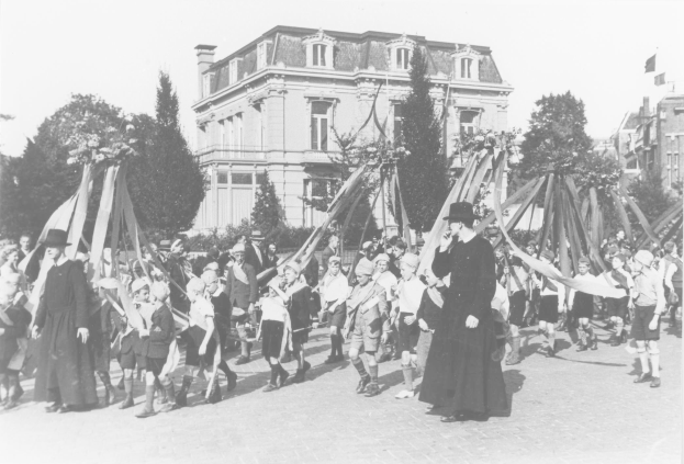 Schwarz-weißes Bild einer Gruppe von Menschen, die eine Straße entlanggehen und Fahnen schwenken, vor einem Gebäude mit Bäumen und einem Maibaum im Hintergrund.
