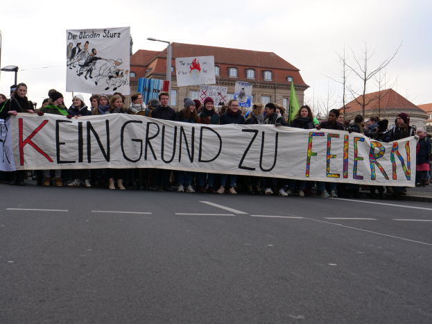Demonstranten mit einem Banner "Kein Grund zu Feiern" gegen deutsche Sparmaßnahmen, im Hintergrund Straßeninfrastruktur und Gebäude unter einem klaren Himmel.