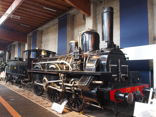 Alte Dampflokomotive in einem Museum ausgestellt mit umgebenen Eisenbahnschienen, Informationsschildern und anderen Ausstellungsstücken, vor einer Wand mit Deckenbeleuchtung.