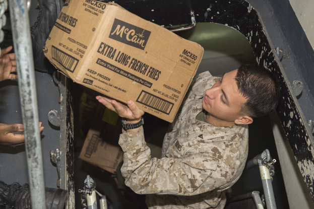 Ein Mann in militärischer Uniform hält eine Kiste mit Lebensmitteln in der Hand, wobei die Hand einer anderen Person zu sehen ist, die die Kiste in einen Lastwagen lädt.