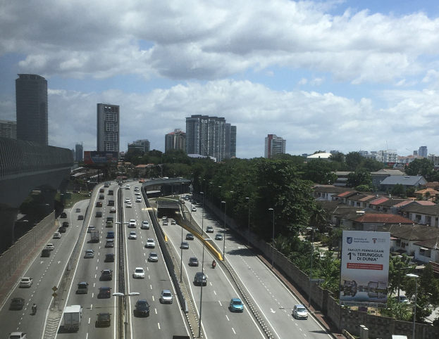 Eine belebte Autobahn mit hohen Gebäuden, eine Brücke auf der linken Seite, Laternenpfähle, Bäume und ein bewölkter Himmel im Hintergrund.
