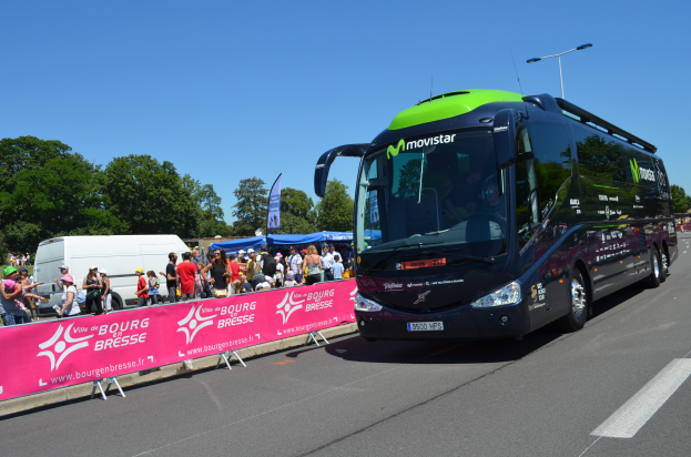 Ein schwarzer und grüner Bus fährt auf einer Straße neben einer Menge Menschen, einige tragen Mönen, mit einer Fahne auf der linken Seite und Bäumen unter einem klaren blauen Himmel im Hintergrund.