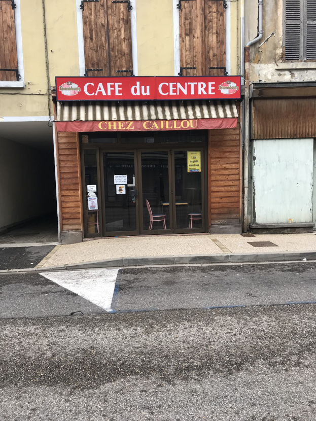 Ein zweistöckiges Gebäude mit einer 'Café du Centre'-Schilder an der Seite, mit Fenstern, einer Tür und einer Straße davor mit einer kleinen Informationsanzeige.