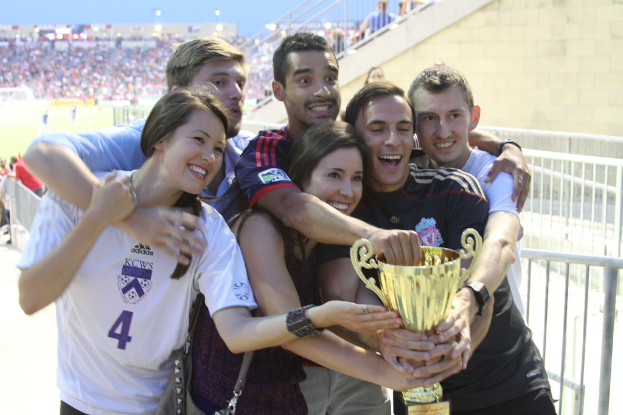 Sechs Personen, darunter zwei Frauen und vier Männer, halten einen Pokal und lächeln, posieren vor einem Eisenzaun, mit einer Wand und einem Zaun oben rechts, Menschen und Lichter im Hintergrund, möglicherweise in einem Stadion.