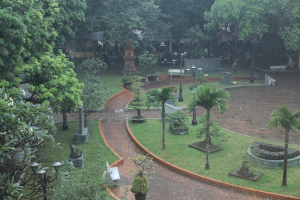 Ein grüner Park mit Bäumen, Pflanzen, Gras, Laternen, Bänken und Skulpturen, mit einem Gebäude im Hintergrund.