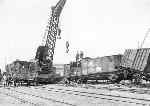 Schwarzes und weißes Bild einer Zugkatastrophe mit einem Kran, der ein Waggon auf die Schienen hebt, umgeben von mehreren Menschen, mit Gebäuden und einem klaren Himmel im Hintergrund.