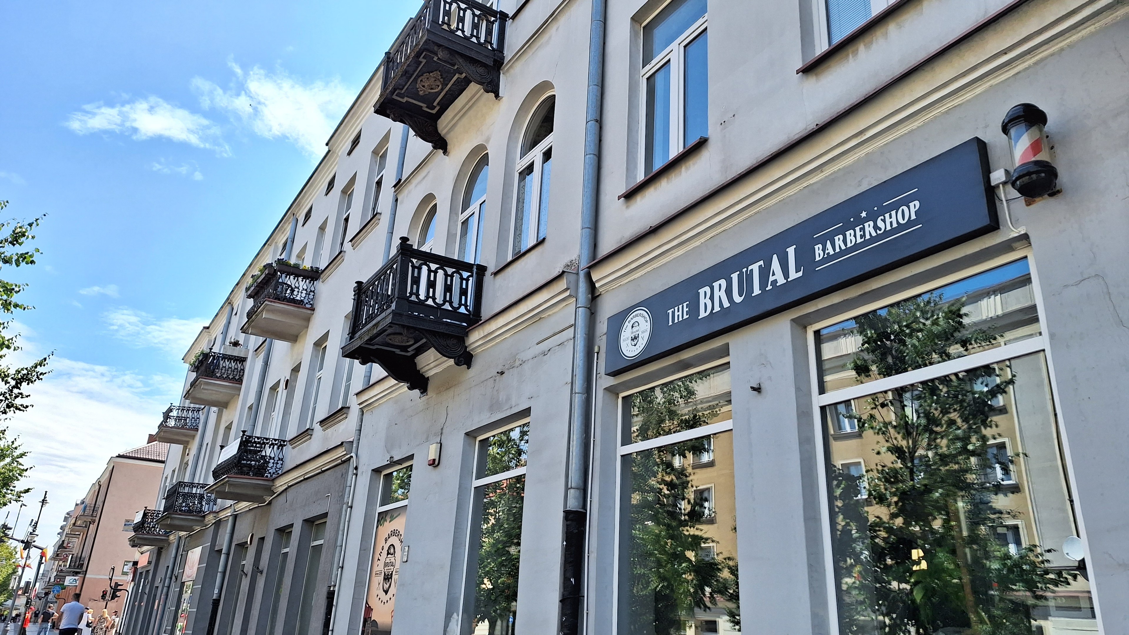 Brutalistischer Barbershop-Außenbereich in Berlin mit eckigen Betongebäuden, Fenstern, Balkonen und Namensschildern, umgeben von Bäumen, Straßenlaternen und Fußgängern unter einem bewölkten Himmel.