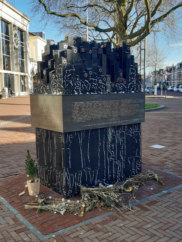 Denkmal in einem Stadtplatz für Holocaust-Opfer mit einem Baum davor, einem Topfgewächs auf dem Boden, verstreuten trockenen Blättern, umgeben von Gebäuden, Fahrzeugen, Pfählen, Bäumen und Himmel.