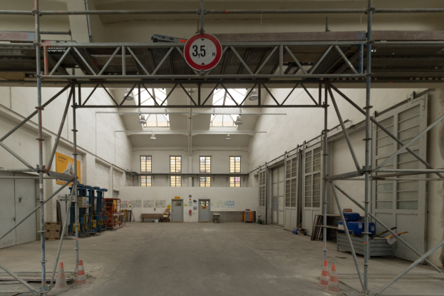 Großes Lagerinnere mit ausgedehnten Gerüsten, Verkehrskegeln, Metallstangen, einem Schild, Maschinen, einer Tür, Fenstern, Wänden, Deckenleuchten und einem Dach.