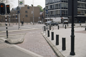Eine Straßenszene in der Stadt mit einem geparkten Fahrrad, fahrenden Fahrzeugen, Fußgängern, Pfählen, Verkehrszeichen, Schildern, Bäumen, Gebäuden mit Fenstern und einem klaren blauen Himmel.