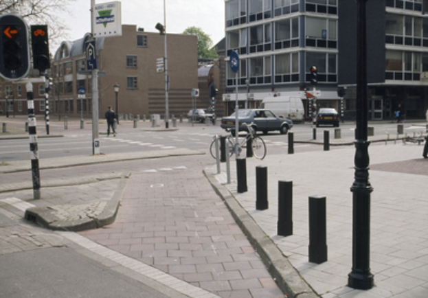 Eine Straßenszene in der Stadt mit einem geparkten Fahrrad, fahrenden Fahrzeugen, Fußgängern, Pfählen, Verkehrszeichen, Schildern, Bäumen, Gebäuden mit Fenstern und einem klaren blauen Himmel.