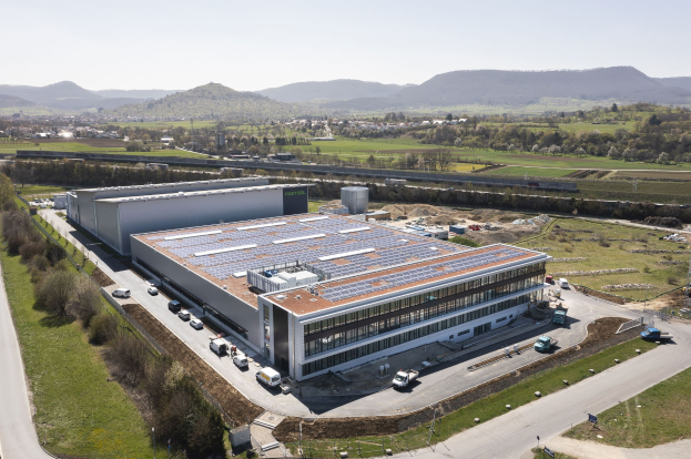 Luftaufnahme eines großen Industriegebäudes mit Solarmodulen auf dem Dach, umgeben von Fahrzeugen, Bäumen, Gras, Strommasten und Leitungen, mit Hügeln und einem klaren blauen Himmel im Hintergrund.
